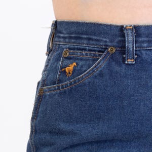 Puede incluir: Primer plano de unos vaqueros azules con un bolsillo con un caballo dorado bordado. Los vaqueros tienen un estilo cl&aacute;sico de cinco bolsillos con remaches y costuras de color lat&oacute;n. Los vaqueros son de lavado medio.