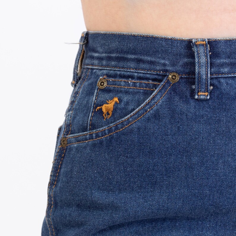 Puede incluir: Primer plano de unos vaqueros azules con un bolsillo con un caballo dorado bordado. Los vaqueros tienen un estilo cl&aacute;sico de cinco bolsillos con remaches y costuras de color lat&oacute;n. Los vaqueros son de lavado medio.