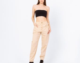 May include: A woman wearing a black tube top and tan pants with gold buttons. The pants have pockets and are cuffed at the ankles. She is also wearing black boots.