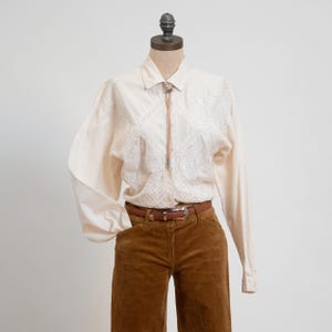 May include: A cream-colored long-sleeve button-down shirt featuring intricate white lace paneling on the front, paired with a brown corduroy belt and a bolo tie with a silver-toned clasp. The shirt has a pointed collar and long sleeves.