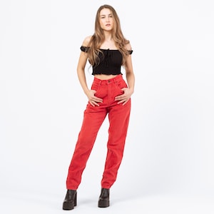 May include: A woman wearing a black off-the-shoulder crop top and high-waisted red jeans. The jeans have a classic five-pocket design. She is wearing black platform boots. The background is plain white.