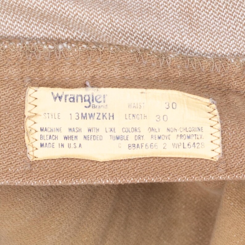 May include: A brown Wrangler brand clothing label with white text. The label states "STYLE 13MWZKH", "WAIST 30", "LENGTH 30", "MACHINE WASH WITH LIKE COLORS ONLY NON-CHLORINE BLEACH WHEN NEEDED TUMBLE DRY REMOVE PROMPTLY", "MADE IN USA", "C 8BAF666 2 WPL6428".