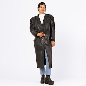 Peut inclure: Un long manteau en cuir noir avec de larges épaulettes et une taille cintrée. Le manteau a un col en V et est porté sur un haut blanc et un jean bleu. Le modèle porte des bottines noires à plateforme.