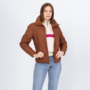 May include: A brown quilted jacket with orange trim and a high collar. The jacket is worn over a cream-colored sweater with a pink stripe. The person is wearing blue jeans.