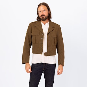 May include: A cropped olive green jacket with four front pockets and button closures. The jacket has a notched collar and a small patch on the left sleeve. The model is wearing a white shirt and dark blue jeans.