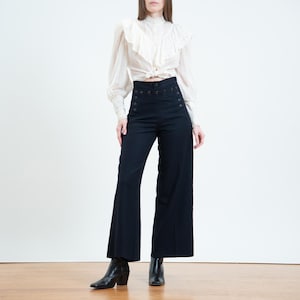 May include: A cream-coloured long-sleeve blouse with a ruffled collar and front button closure, paired with high-waisted black wide-leg trousers featuring a double row of buttons at the waist and black leather ankle boots.
