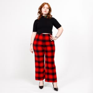 May include: A woman wearing a black mock neck shirt and red and black plaid pants. She is standing with her hand on her hip and looking at the camera.