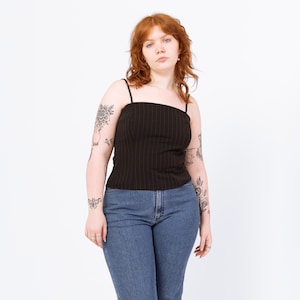 Peut inclure: Un caraco noir à fines rayures verticales et bretelles spaghetti. Le haut est associé à un jean en denim bleu. Le modèle a les cheveux roux et des tatouages sur les bras.