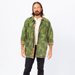 May include: A green camouflage jacket with a leaf pattern. The jacket has a collared neckline, a zipper closure, and two front pockets. The sleeves are three-quarter length, and the cuffs are a brown color. The jacket is worn over a white shirt and dark blue jeans.