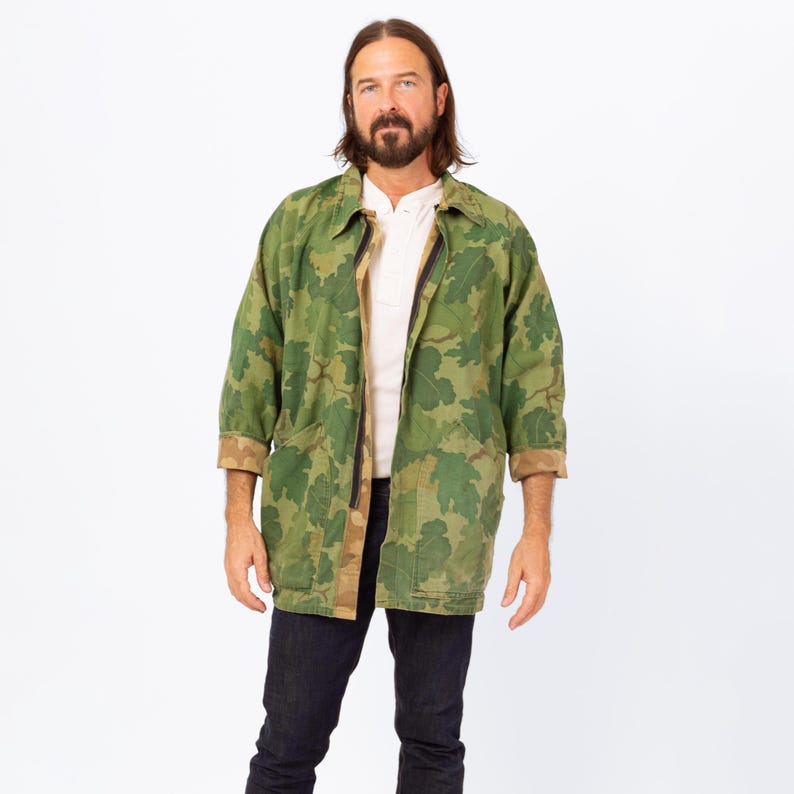 May include: A green camouflage jacket with a leaf pattern. The jacket has a collared neckline, a zipper closure, and two front pockets. The sleeves are three-quarter length, and the cuffs are a brown color. The jacket is worn over a white shirt and dark blue jeans.