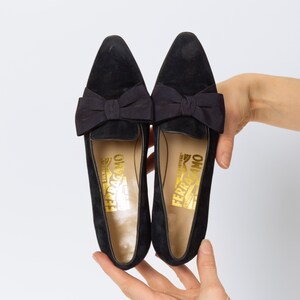 Puede incluir: Un par de mocasines negros de ante con un lazo. Los zapatos tienen una punta afilada y un logotipo dorado en la plantilla. Los mocasines se muestran sobre un fondo blanco.