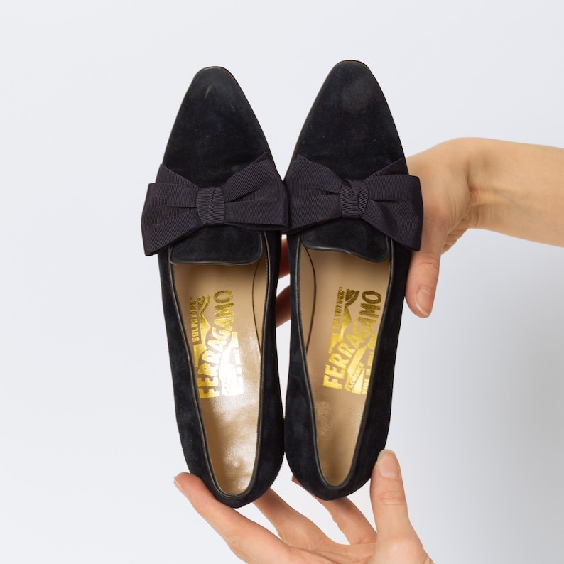 Puede incluir: Un par de mocasines negros de ante con un lazo. Los zapatos tienen una punta afilada y un logotipo dorado en la plantilla. Los mocasines se muestran sobre un fondo blanco.