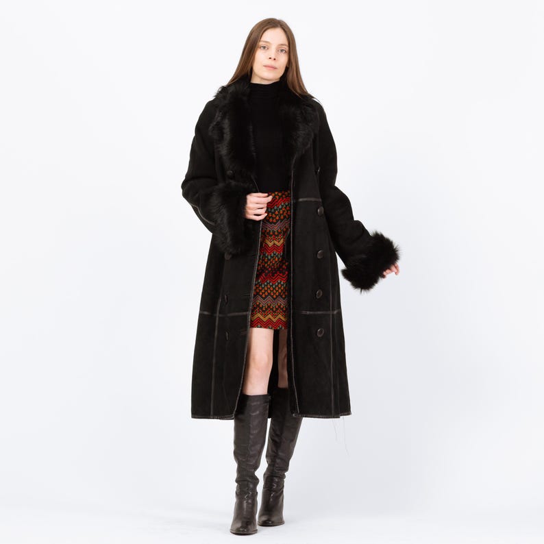 May include: A long black coat with fur trim on the collar and cuffs. The coat has a button-down front and is worn over a black turtleneck and a patterned skirt. The model is wearing knee-high black boots.
