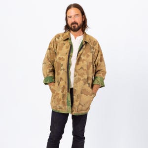 May include: A camouflage-patterned jacket in shades of brown, tan, and green. The jacket has a black zipper, two front pockets, and green cuffs. The person is wearing a white shirt and dark blue jeans.
