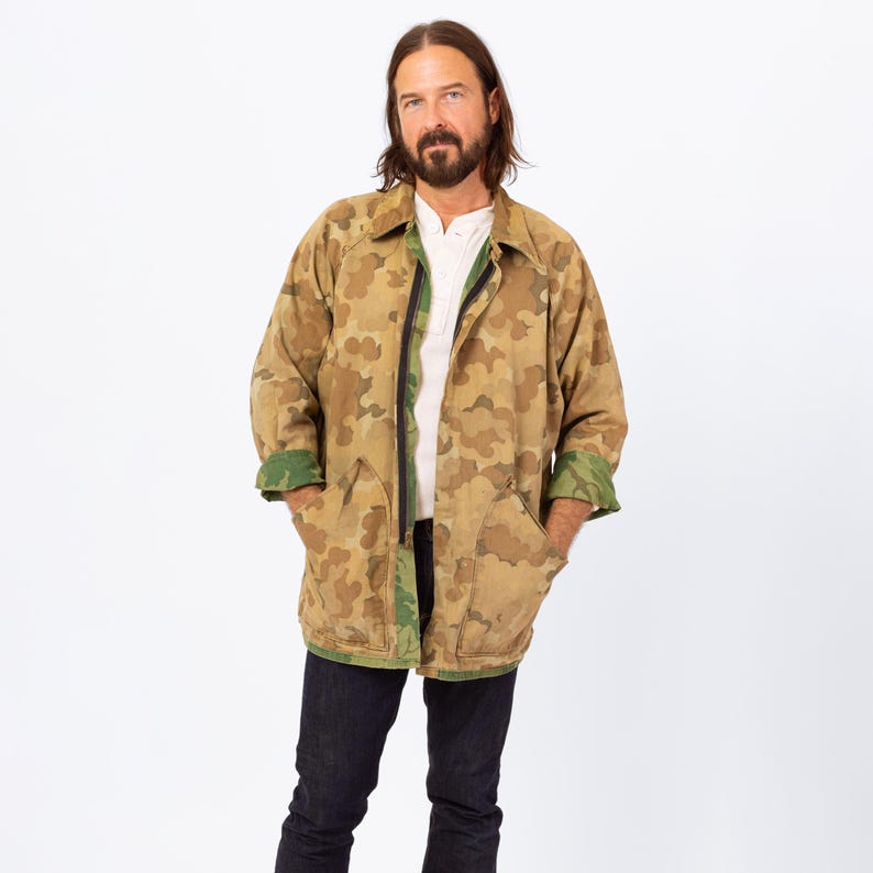 May include: A camouflage-patterned jacket in shades of brown, tan, and green. The jacket has a black zipper, two front pockets, and green cuffs. The person is wearing a white shirt and dark blue jeans.