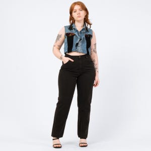May include: A sleeveless denim vest with black panels and floral accents, paired with black high-waisted jeans. The vest has a ruffled collar and decorative details. The jeans are a classic, straight-leg style, and the model is wearing black sandals.