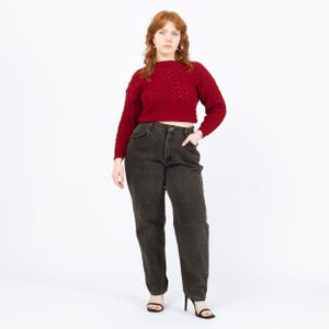 May include: A woman wearing a dark red, textured knit sweater and dark gray denim jeans. The sweater has a crew neck and long sleeves. The jeans are high-waisted and straight-legged. The woman is wearing black heels.