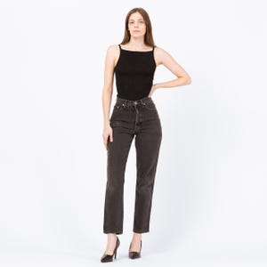 May include: A woman wearing a black tank top and dark wash denim jeans. The jeans have a high waist and a button fly. The woman is also wearing black high heels.