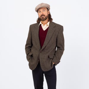 Puede incluir: Una blazer de tweed marrón con un suéter burdeos y una camisa de cuello claro. La blazer tiene dos bolsillos delanteros y un cierre de un solo botón. Una gorra de estilo newsboy estampada completa el look. El pantalón es azul oscuro.