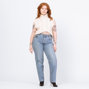 May include: A woman wearing a cream-colored cable knit sleeveless turtleneck sweater and light blue denim jeans. The jeans have a classic straight leg fit. The woman is also wearing black heeled boots.