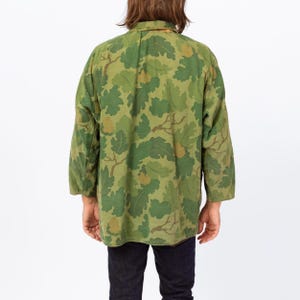 May include: Green camouflage jacket with a leaf pattern. The jacket has long sleeves and a button-down collar. The camouflage design features shades of green and brown. The jacket is worn over dark blue jeans.