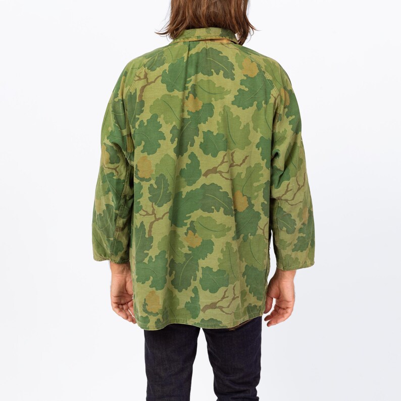 May include: Green camouflage jacket with a leaf pattern. The jacket has long sleeves and a button-down collar. The camouflage design features shades of green and brown. The jacket is worn over dark blue jeans.