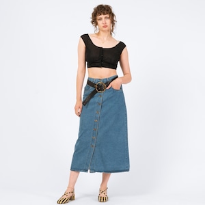 May include: A woman wearing a black crop top and a button-down denim skirt. The skirt is a light blue wash and has a front slit. A black belt with a gold buckle cinches the waist. The woman is also wearing striped sandals.