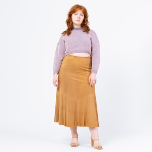 Puede incluir: Una mujer con un suéter morado claro de canalé y una falda midi de ante color canela con dobladillo acampanado. Lleva tacones altos color canela. El conjunto está sobre un fondo blanco.