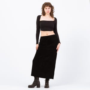 May include: A woman wearing a black long-sleeved crop top and a black corduroy maxi skirt. She is also wearing black ankle boots with a block heel. The outfit is set against a white background.