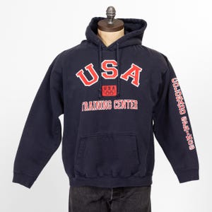 Puede incluir: Una sudadera con capucha azul marino con el texto "USA" en letras rojas y blancas, "USA" en un pequeño cuadro con los anillos olímpicos, y "TRAINING CENTER" en letras rojas. La manga derecha tiene el texto "COLORADO SPRINGS" en letras rojas.