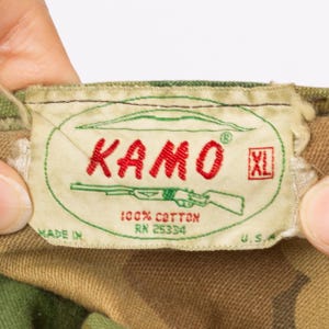 May include: Close-up of a tag with the word "KAMO" in red, a green outline, and a rifle illustration. The tag also includes the text "100% Cotton" and "XL" in a red box. The tag is on a camouflage fabric.