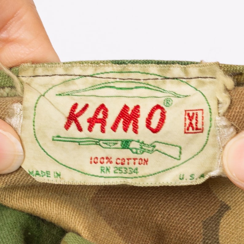 May include: Close-up of a tag with the word "KAMO" in red, a green outline, and a rifle illustration. The tag also includes the text "100% Cotton" and "XL" in a red box. The tag is on a camouflage fabric.