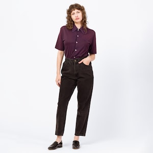 Peut inclure: Une personne porte une chemise boutonnée à manches courtes, violet foncé, et un jean marron foncé. La chemise a un col et une seule poche. Le jean est taille haute et droit. La personne porte des mocassins noirs.