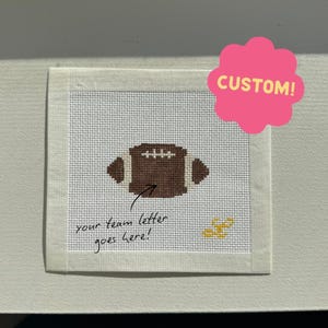 May include: A square needlepoint canvas featuring a brown football design with white laces. The canvas includes the handwritten text 'your team letter goes here!' pointing to the football, a small yellow stitched detail, and a pink 'CUSTOM!' sticker.