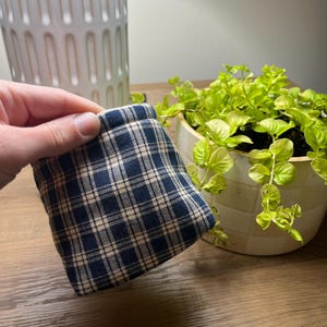 Puede incluir: Pequeño objeto de tela cuadrado con un patrón de cuadros azul marino y beige. El objeto se sostiene en una mano, con un jarrón blanco y una planta en maceta en el fondo. La tela parece ser de algodón o lino suave.