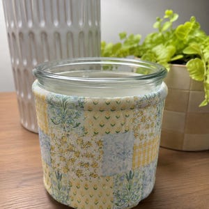 May include: A clear glass jar wrapped in a quilted fabric sleeve featuring a patchwork pattern of yellow and light blue floral, gingham, and botanical prints. The fabric cover is stitched with a soft texture and fits snugly around the cylindrical glass container.