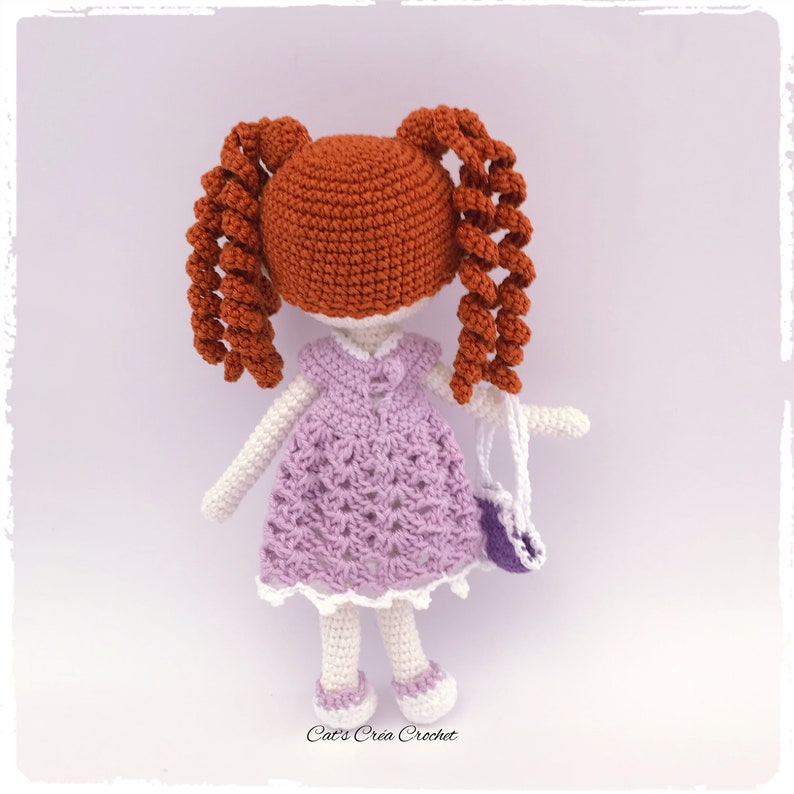 Op de afbeelding: Een gehaakte pop met rood haar in vlechtjes, die een lavendelkleurige jurk met witte rand draagt en een paarse handtas draagt. De pop staat op een witte achtergrond.