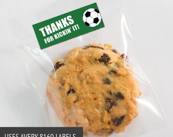 May include: A clear cellophane bag containing a large, golden-brown cookie with chocolate chips and raisins. A green label with the text "THANKS FOR KICKIN' IT!" and a soccer ball graphic is affixed to the bag.