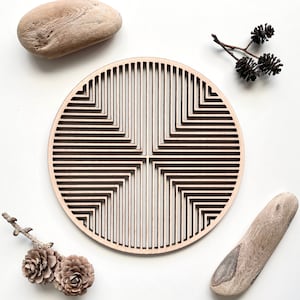 Könnte beinhalten: Eine runde Holz-Wanddekoration mit einem geometrischen Muster aus sich kreuzenden Linien. Die Dekoration ist hellbraun und hat eine natürliche Holzoptik. Sie ist umgeben von kleinen Stücken Treibholz und Tannenzapfen.