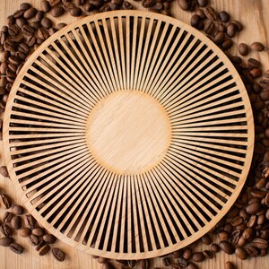 Peut inclure: Un plateau circulaire en bois présentant un motif en rayons de soleil composé de fines lattes rayonnantes entourant un disque central plein. Le plateau en bois clair est entouré de grains de café torréfiés brun foncé.