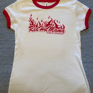 May include: A white ringer t-shirt with red trim on the collar and sleeve cuffs. The front features a red graphic of flames above the text "Red Hot Mama" and "she's SMOKIN'!" in a stylized red font. The shirt has short sleeves and a crew neckline.