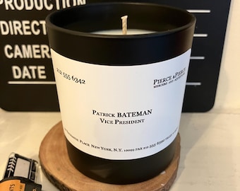 May include: A black candle in a cylindrical glass with a white label featuring the text "Patrick Bateman, Vice President." The candle sits on a wooden coaster, with film strips and a Hollywood clapperboard in the background.
