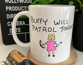 May include: White ceramic mug with the text "Buffy Will Patrol Tonight" and a cartoon illustration of a blonde girl in a pink dress holding a bow and arrow. The mug sits on a wooden coaster, with film reel and a clapperboard in the background.
