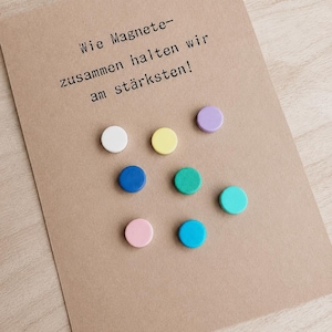 Può includere: Un biglietto di carta kraft con testo in tedesco e sette magneti rotondi colorati. I magneti sono bianchi, gialli, viola, blu, verdi, rosa e turchese. Il testo recita "Wie Magnete - zusammen halten wir am stärksten!"