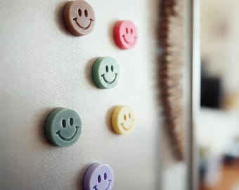 May include: A selection of colourful smiley face magnets in shades of brown, pink, green, yellow, and purple. The round magnets are attached to a silver fridge door. The smiley faces have a simple, classic design.