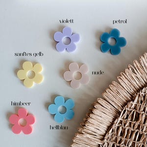 May include: Six flat, flower-shaped acrylic charms with a central circular cutout. The colors include violet, petrol blue, soft yellow, nude, raspberry pink, and light blue. Each flower is labeled with its respective color name in text.