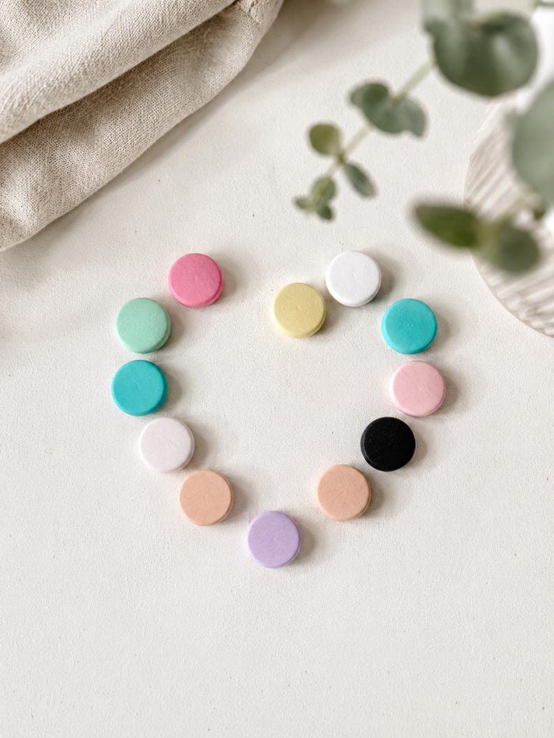 May include: A collection of twelve small, circular, flat wooden discs arranged in a heart shape. The discs feature a variety of solid matte colours including pink, mint green, teal, white, yellow, black, peach, lavender, and light pink.