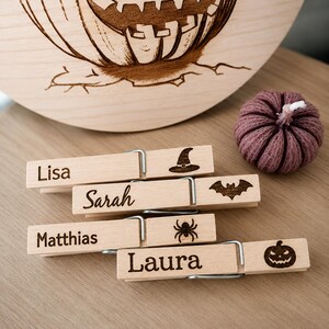 Könnte beinhalten: Vier helle Holzwäscheklammern, graviert mit den Namen Lisa, Sarah, Matthias und Laura, jeweils mit einem individuellen Halloween-Motiv wie Hexenhut, Fledermaus, Spinne und Kürbisgesicht, ergänzt durch einen kleinen violetten Kürbis aus Stoff.