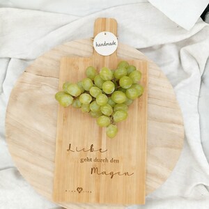 Puede incluir: Tabla de cortar de madera con uvas verdes. La tabla tiene grabada la frase "Liebe geht durch den Magen". Una pequeña etiqueta que dice "handmade" está adjunta. La tabla está sobre una superficie redonda de madera.