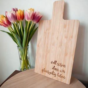 Może przedstawiać: Jasna drewniana deska do krojenia z uchwytem, z wygrawerowanym tekstem. Bukiet tulipanów w szklanym wazonie znajduje się po lewej stronie. Tulipany mają żółte, pomarańczowe i różowe płatki.